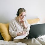A focused woman in glasses and headphones works on a laptop from a cozy bed.